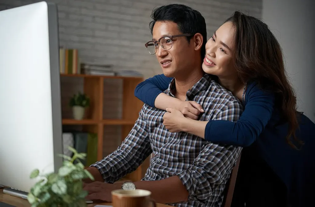 A wife hugging her husband from the back, looking at contractors online
